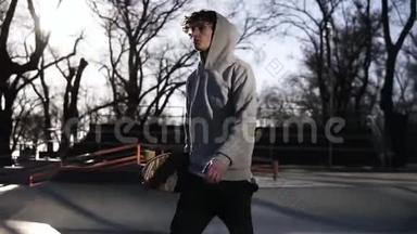 穿着灰色连帽衫的年轻滑板手拿着滑板在滑板公园里散步。 侧视。 慢动作