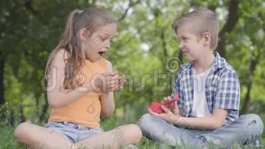漂亮的小女孩和坐在草地上的英<strong>俊男</strong>孩在一起。 <strong>男</strong>孩拿着一个红色的小盒子打开它