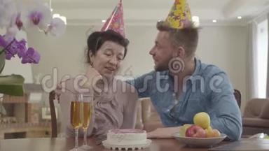 肖像成熟的女人和成年的孙子坐在<strong>桌子</strong>旁，头上戴着生日帽。 在<strong>桌子</strong>上