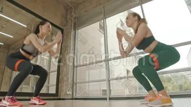 两个健康的女人站在工作室里，用大腿之间的拉伸皮带训练她们的腿-蹲着
