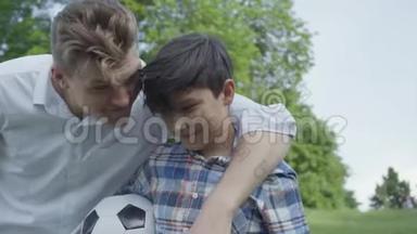 那个孩子在公园的前台拿着足球。 年轻的爸爸过来拥抱男孩，看着，笑着走进