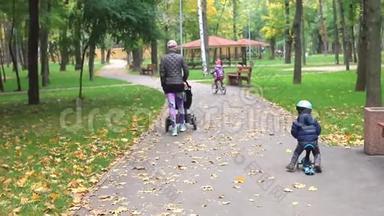 小男孩骑着滑板车，骑着平衡车，骑在柏油路旁，妈妈在城市公园附近散步。