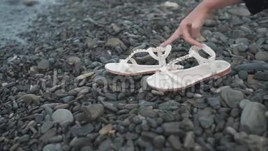 白色凉鞋在岩石和潮湿的海滨。 女人`手拿凉鞋。