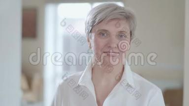一幅漂亮的穿着白色衬衫的高级女人的肖像，在镜头里看着，在室内靠近时露出了愉快的微笑。 背景