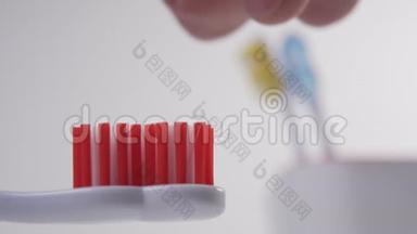 一只手把一支蓝色牙膏挤在一支红色刚毛的白色塑料牙刷上，贴在一个有黄色和蓝色牙刷的玻璃杯上