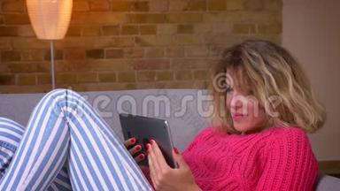 近景拍摄的金发家庭主妇穿着<strong>粉红</strong>色毛衣，躺在沙发上，在舒适的家庭氛围中，在平板电脑上用<strong>视频</strong>聊天。