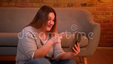 超重的女模特坐在地板上，在平板电脑上积极地用视频聊天，在舒适的家庭氛围中欢笑。