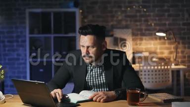 一个留胡子的人晚上在办公室用笔记本电脑，在办公桌上用笔记本做笔记