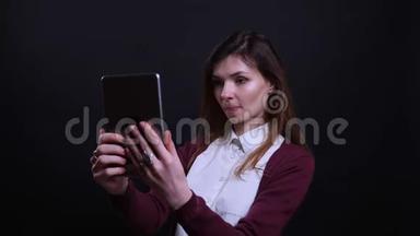 年轻的黑发女商人在黑色背景下愉快地在平板电脑上制作漂亮的自拍照片的肖像。