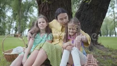 肖像可爱的成熟女人亲吻她的两个可爱的孙女，坐在公园树下的草地上，看着
