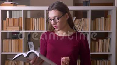 年轻漂亮的黑发女学生戴着眼镜，在<strong>图书馆</strong>里<strong>看书</strong>，看着镜头。