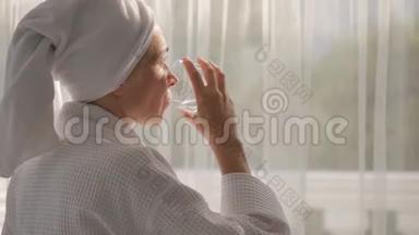 一个穿着<strong>白大褂</strong>，头上拿着毛巾的女人，从站在大窗户边的玻璃杯里喝水..