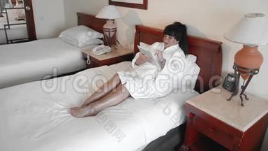 一名白人妇女穿着白色浴袍躺在酒店的床上，用平板电脑上网。概念