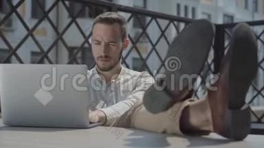 肖像胡子的年轻人坐在露<strong>台</strong>上的<strong>桌</strong>子上，双脚放在<strong>桌</strong>子上，笔记本电脑躺在附近。 概念