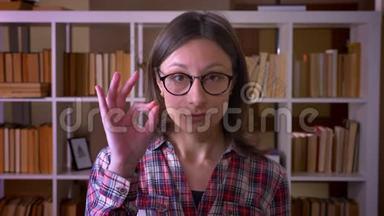 一位戴眼镜的年轻有魅力女学生的特写镜头，在室内的图书馆里看着摄像机，显示出一个好的标志