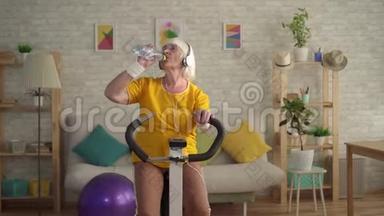 戴耳机的运动老妇人在运动自行车上进行有氧训练，从瓶子里喝水