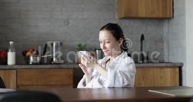 一个穿着白色浴袍的成年女人坐在厨房里，手里拿着手机..
