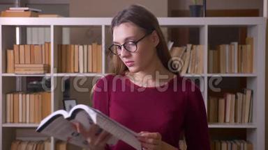 年轻漂亮的黑发女学生戴着眼镜，看书，在图书馆里微笑着相机。