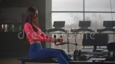 一个坐着的女人在健身房的大窗户和跑步机的背景下拉动模拟器的重量