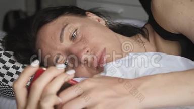 一个幸福的女人躺在她可爱的卧室里，躺在白色的床上，在智能手机上发短信。 技术、互联网