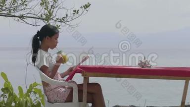在海滩咖啡馆的桌旁看书时喝果汁的少女