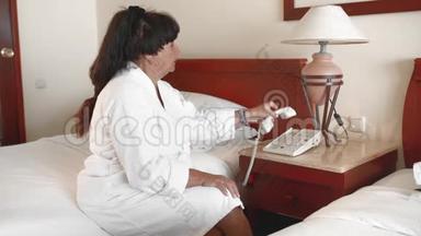 一位不快乐的老太太穿着白色浴袍坐在酒店的床上打电话。慢动作