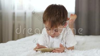 一个可爱的小男孩躺在床上看着智能手机屏幕。 孩子在智能手机上玩。
