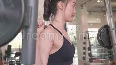 漂亮的亚洲女孩穿着运动服在<strong>健身</strong>运动<strong>健身</strong>房举起杠铃。 运动员生活方式侧视图