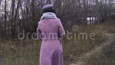 秋天，女人走在一条杂草丛生的小路上。 库存录像。 穿着外套的漂亮年轻女子在高大的人群中漫步