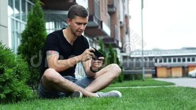 年轻人在智能手机上玩手机游戏，坐在大楼附近的草地上。