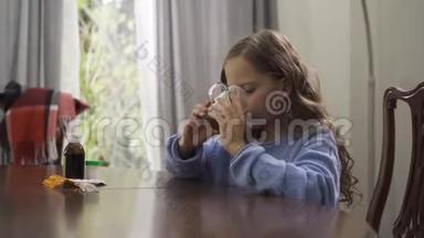 可爱的小女孩一边喝凉茶，一边坐在家里的桌子上。 在厨房里喝茶治<strong>感冒</strong>的孩子