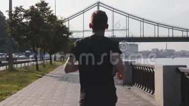 年轻苗条男子在城市码头沿河奔跑的背面景色。 肌肉活跃的男孩跑得很快。 运动员沿着河跑