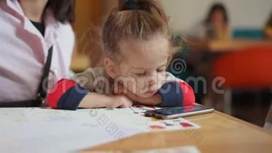 一个欧洲貌美的小女孩正坐在<strong>妈妈怀里</strong>的一张桌子上，看着智能手机上的视频