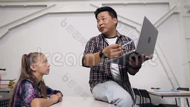 年轻的韩国老师坐在桌子上，手里拿着笔记本电脑，给学生们上课。 小学学生