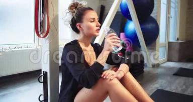 口渴的女士在健身房辛苦锻炼了一天后，从塑料瓶里喝了些水，她坐在地板上