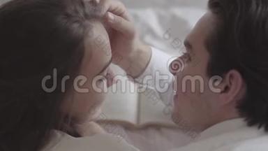 一对幸福的年轻夫妇躺在床上看书特写。 英俊的男人摸着女人的脸。 投标