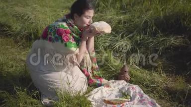 美丽的超重女人坐在野草中，拿着面包和土罐准备吃。 传统