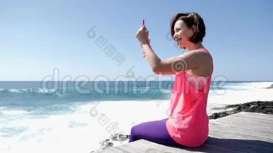 年轻美丽的白种人女人坐在<strong>岩石</strong>海滩上拿着电话，拍着<strong>海浪</strong>拍打<strong>岩石</strong>的照片