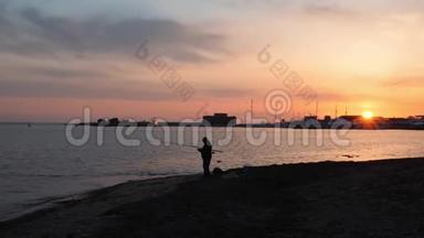 孤独男人的剪影，带着钓鱼竿站在空荡荡的海滩上，地平线上有粉红色的日落。 日落时分钓鱼人剪影