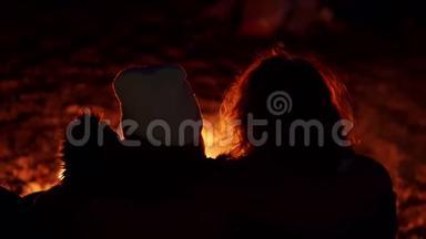 一对年轻夫妇坐在火炉旁。 在海边用帐篷露营。 两个视频的序列。 手持设备的背面视图