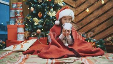 4k<strong>视频</strong>可爱的微笑女孩坐在舒适的家里，喝热巧克力在<strong>圣诞树</strong>上的木制背景。