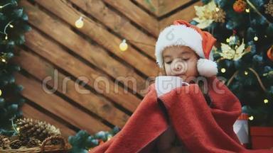 4k视频可爱的微笑女孩<strong>坐在</strong>舒适的家里，喝热巧克力在圣诞<strong>树上</strong>的木制背景。