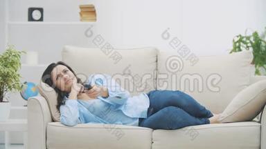 年轻，放松的女人正在用Zapper切换频道。 她正在看电视，躺在家里的沙发上。 复制空间。 4K. 慢慢