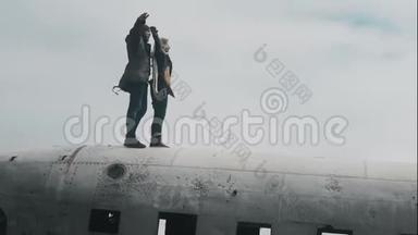 一对年轻的旅行夫妇站在冰岛坠毁的DC-3飞机上，在大风天一起举手。