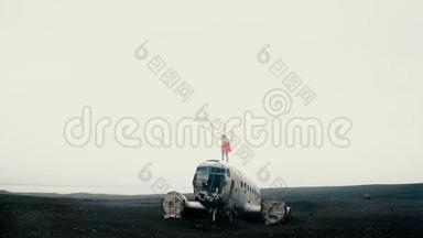 年轻的旅行妇女站在冰岛坠毁的DC-3飞机的顶部，在刮风的日子脱下粉红色的夹克。