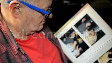 高加索族的老人，戴着蓝色的眼镜，带着照片翻阅相册，坐着。 记忆和概念