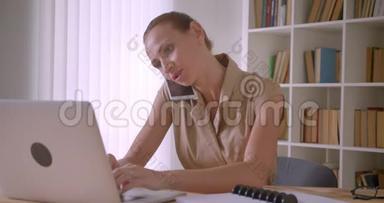 一位优雅的白人女商人正在分析笔记本电脑上的数据，她在办公室里<strong>忙着</strong>用手机交谈，在笔记本上写字。