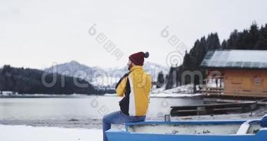 在一个白雪皑皑的湖泊和<strong>森林</strong>旁边，一位魅力非凡的年轻<strong>游客</strong>欣赏他坐在一艘蓝色小船旁边的景色。