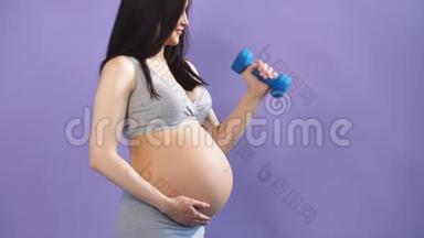 快乐的怀孕妈妈在紫罗兰背景的哑铃工作室锻炼