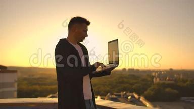 镜头跟随着一个人在日落时分带着笔记本电脑和一个瓶子穿过屋顶。 自由职业者打开笔记本电脑